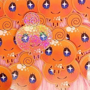 May include: A collection of orange stickers with a smiling, cartoonish ghost design. The ghost has blue and white eyes, a curly top, and a dripping bottom. One sticker is covered in glitter.