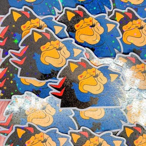 May include: A collection of die-cut stickers featuring a stylized character with black and blue fur, yellow ears, and a tan muzzle. The design includes red accents and a holographic finish with small star-shaped sparkles across the surface.