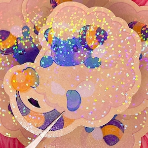 May include: A collection of glittery stickers featuring a cartoon sheep with a fluffy, cloud-like body. The sheep has blue, purple, and orange accents. The stickers have a holographic effect, with a light pink background.