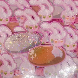 Könnte beinhalten: Eine Sammlung von Aufklebern mit einer Cartoon-Figur mit einem hasenähnlichen Kopf und einem schneckenartigen Körper. Die Aufkleber sind überwiegend rosa und weiß, mit Glitzerakzenten und einem niedlichen, skurrilen Design.