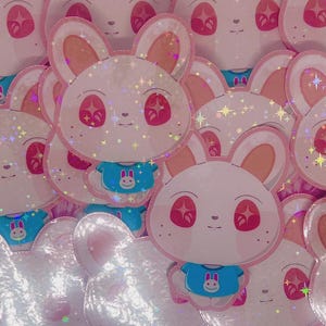 Puede incluir: Una colección de pegatinas con un conejo de dibujos animados con pelaje blanco, orejas rosas y ojos rojos. El conejo lleva una camiseta azul con un gráfico de conejo. Las pegatinas tienen un acabado holográfico y brillante.