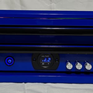 Puede incluir: Una caja azul con listones horizontales negros y perillas plateadas. Tiene una pantalla digital que dice "VOLTAGE 2.2 DC V" y un botón de encendido azul. La caja está sobre una superficie blanca.