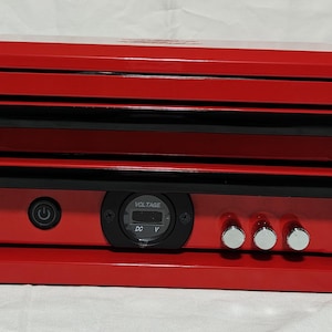 Puede incluir: Una caja roja con detalles negros, que incluye un botón de encendido, un voltímetro y tres perillas plateadas. La caja tiene una tapa con bisagras y varias ranuras horizontales. El diseño es limpio y moderno.