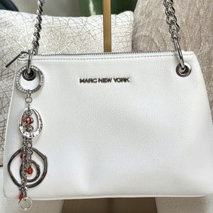 May include: White handbag with silver chain straps and a decorative charm. The bag features the text "MARC NEW YORK" and a zipper closure. The charm includes a ring of rhinestones, an oval pendant, and a circular pendant with orange beads.