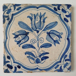 May include: A square ceramic tile featuring a hand-painted blue floral motif on an off-white background. The design displays a central flower flanked by two smaller blossoms, all enclosed within a decorative blue border with corner accents.