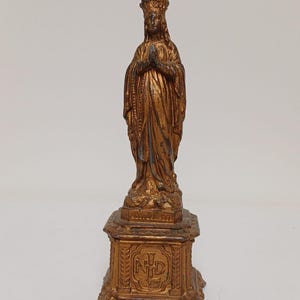 Könnte beinhalten: Goldfarbene Statue einer Frau mit Krone, die Hände zum Gebet gefaltet, auf einem dekorativen Sockel stehend. Die Statue weist aufwendige Details auf und ist wahrscheinlich aus Metall gefertigt. Der Sockel hat Text und ein dekoratives Design.