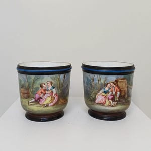 May include: Two antique porcelain planters with hand-painted scenes of couples in a garden setting. Each planter has a black base, a blue rim, and a white interior. The planters are likely from the 19th century.