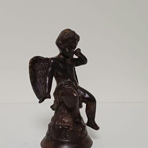 Könnte beinhalten: Eine bronzefarbene Cherub-Statue mit detaillierten Flügeln, die auf einem strukturierten Sockel sitzt. Der Cherub ist mit einer Hand am Gesicht dargestellt, was auf Kontemplation hindeutet. Die Statue ist ein dekorativer Gegenstand.