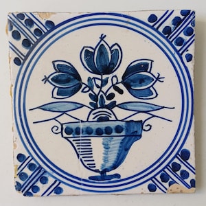 Op de afbeelding: Een vierkante keramische tegel met een handgeschilderd blauw-wit ontwerp. Het midden toont een bloemstuk in een decoratieve pot, omsloten door een cirkelvormige rand. De hoeken zijn versierd met blauwe stippenpatronen op een witte achtergrond.