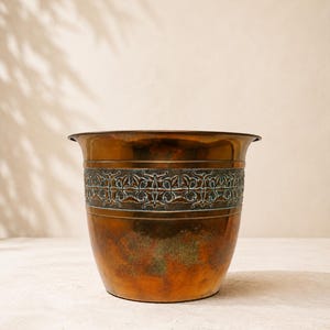 May include: A round copper planter featuring a wide, embossed band around the upper section with a dark, oxidized floral pattern. The lower body displays a mottled, weathered patina with varying shades of brown, orange, and green metallic tones.