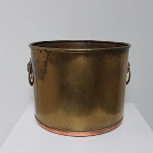 Peut inclure: Un seau cylindrique en laiton avec un bord de base en cuivre poli. Le récipient présente deux poignées latérales en forme de têtes de lion, chacune tenant un anneau circulaire. La surface en laiton présente une patine vieillie avec des signes d'oxydation.