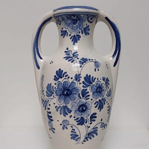 May include: A white ceramic vase with two handles and a blue floral design. The vase features a detailed pattern of blue flowers and leaves, with blue accents on the rim and handles. The vase is approximately 30 cm tall.