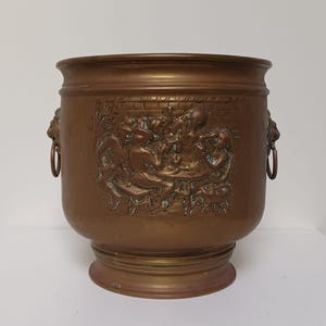 May include: A cylindrical brass bucket featuring a relief scene of figures gathered around a table. The vessel has a wide rim, a pedestal base, and two side handles shaped like lion heads with attached rings, displaying a warm metallic finish.