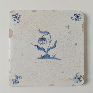 May include: A square ceramic tile with a white base and a visible network of fine cracks across the surface. The center features a blue hand-painted illustration of a single flower with leaves, while each corner displays a small blue floral motif.