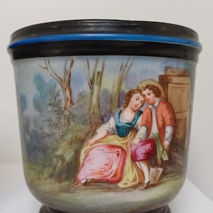 May include: A decorative ceramic container with a black rim and base, featuring a painted scene of a couple in a garden. The couple is dressed in period clothing, with the woman in a pink dress and the man in a red jacket. The container has a blue accent.