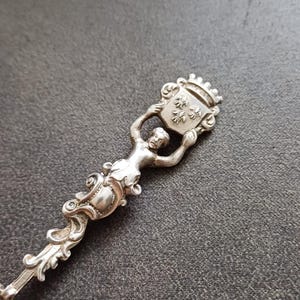 May include: A silver-toned metal spoon handle featuring a sculpted figure holding a shield above its head. The shield is topped with a crown and displays three fleur-de-lis symbols. The lower portion of the handle has ornate, swirling scrollwork.