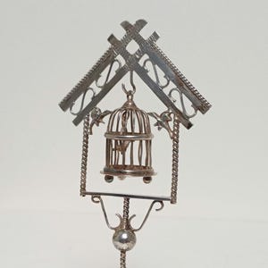 Peut inclure: Un ornement décoratif en métal argenté composé d'un cadre en forme de maison avec un toit, des supports latéraux en métal torsadé et une petite cage à oiseaux suspendue à un crochet central. La cage contient une figurine d'oiseau et l'ensemble est monté sur une tige métallique fine et torsadée avec une base sphérique.