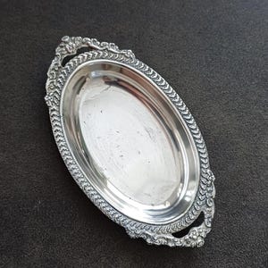 Op de afbeelding: Een ovaal zilverkleurig dienblad met een gepolijste, reflecterende kom. De rand is voorzien van een ingewikkeld, verhoogd reliëfpatroon van bladeren en bloemmotieven, met decoratieve handgrepen aan beide uiteinden van het dienblad.
