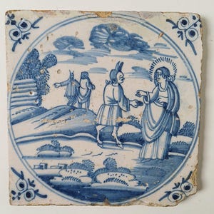 Puede incluir: Un azulejo de cerámica cuadrado con una escena pintada a mano en azul y blanco dentro de un borde circular. La ilustración muestra a una figura con cuernos y pezuñas ofreciendo un objeto a una persona con halo, con otras dos figuras en el paisaje.