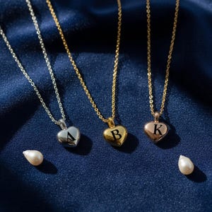 May include: Three heart-shaped pendant necklaces with initial letters A, B, and K. The pendants are silver, gold, and rose gold, suspended on delicate chains. Two pearls are placed near the necklaces on a dark blue fabric.