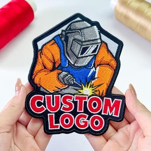 May include: An embroidered patch featuring a welder wearing a gray helmet and orange protective gear, working on a project with a bright yellow spark. The text CUSTOM LOGO is stitched in bold red letters with a white outline at the bottom of the patch.