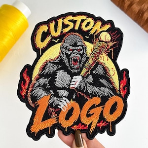 May include: An embroidered patch featuring a black gorilla with red eyes, holding a baseball bat wrapped in barbed wire. The text CUSTOM is stitched in yellow above the gorilla, and the word LOGO is stitched in orange below, with flames.