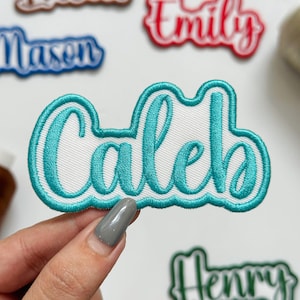 May include: A custom embroidered name patch featuring the name Caleb in a teal cursive script font. The lettering is outlined in a thick teal border against a white fabric background, creating a raised, textured appearance for the name.