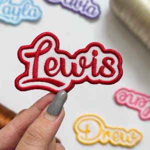 May include: A custom embroidered name patch featuring the name Lewis in red cursive script with a white border. The patch has a thick red stitched outline and a white fabric background. Other similar name patches are visible in the background.