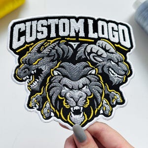 May include: An embroidered patch featuring the text CUSTOM LOGO in white letters at the top. Below the text are three detailed creature heads in shades of gray and black with yellow outlines, including a dragon, a ram, and a central lion.