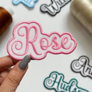 May include: An embroidered patch featuring the name Rose in pink cursive script lettering. The text is set against a white background with a pink stitched border, creating a cloud-like shape. The patch is held up against a white surface.