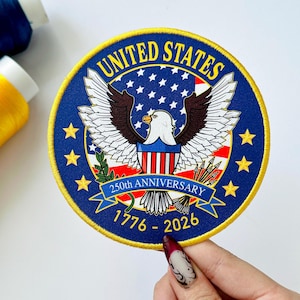 May include: A round, embroidered patch with a blue background and a gold border. The patch features a white eagle with outstretched wings, a red and white striped shield, and the text "UNITED STATES" and "250th ANNIVERSARY 1776-2026".