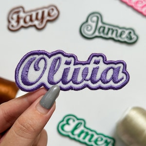 May include: An embroidered patch featuring the name Olivia in purple cursive script with a white outline and a purple border. Other name patches including Faye, James, and Elmer are visible in the background with similar designs.