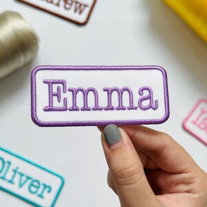 May include: A rectangular embroidered patch with a white background and a purple border. The name Emma is stitched in the center in purple thread using a serif font. The patch has a clean, finished edge and is designed for fabric application.