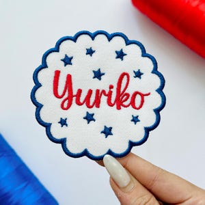 May include: A circular embroidered patch featuring the name "Yuriko" in red script, surrounded by blue stars and a scalloped blue border. The patch is white and appears to be made of fabric.