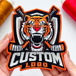 May include: An embroidered patch featuring a snarling tiger head with orange, black, and white fur, positioned above two crossed hockey sticks. The word CUSTOM is stitched in large white letters above the word LOGO in orange, all on a black background.