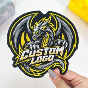 May include: A circular embroidered patch featuring a stylized dragon in black, silver, and yellow thread. The dragon is depicted with wings spread and claws extended. The text CUSTOM LOGO is embroidered in white and yellow across the center.