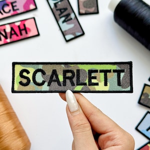 May include: Embroidered patch with the name "SCARLETT" in black letters on a camouflage background. The patch is rectangular with a black border. Other patches with names are visible.