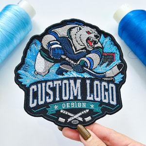 May include: An embroidered patch featuring a white polar bear wearing a dark blue hockey jersey and holding a hockey stick. The patch includes the text CUSTOM LOGO DESIGN in white and blue lettering, with two crossed hockey sticks at the bottom.