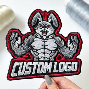 May include: An embroidered patch featuring a muscular, grey rabbit character with red eyes and sharp teeth, posed with flexed arms. The design is outlined in black and red thread, with the text CUSTOM LOGO in white block letters at the bottom.