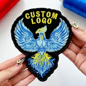 May include: Embroidered patch featuring a blue and yellow phoenix design with outstretched wings on a black background. The text "CUSTOM LOGO" is embroidered in yellow above the bird. The patch is held by a person.