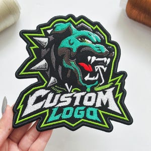 May include: An embroidered patch featuring a stylized snarling wolf head in teal, black, and white with silver spikes. Below the wolf, the text CUSTOM LOGO is stitched in white and teal letters against a black background with a bright green border.