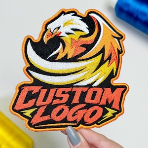 May include: An embroidered patch featuring a stylized eagle head with white, yellow, and orange feathers against a black background. Below the eagle, the text CUSTOM LOGO is stitched in bold orange letters with a thick orange border outline.