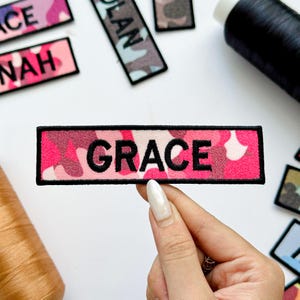 May include: A rectangular patch with the name "GRACE" embroidered in black on a pink camouflage pattern background. The patch has a black border. Other patches with names are visible.