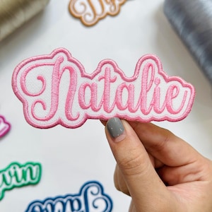 May include: An embroidered fabric patch featuring the name Natalie in a pink cursive script font with a white background and a pink stitched border. The patch has a scalloped edge design that follows the shape of the lettering.
