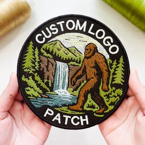 May include: A circular embroidered patch featuring a brown Bigfoot figure walking through a forest landscape with a waterfall and mountains. The text CUSTOM LOGO is arched at the top and the word PATCH is at the bottom in white thread.