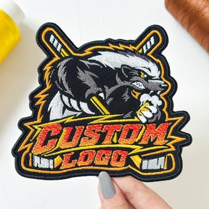 May include: An embroidered patch featuring a stylized badger mascot in black, white, and gray, positioned in front of two crossed hockey sticks. The text CUSTOM LOGO is stitched in red and orange across the bottom, framed by yellow lightning bolts.
