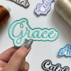 May include: An embroidered patch featuring the name Grace in a teal cursive script font. The lettering is outlined in a matching teal thread and set against a white, ribbed fabric background with a scalloped, rounded border.