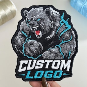 May include: An embroidered patch featuring a snarling bear with grey fur, glowing blue eyes, and a red mouth. The bear is surrounded by bright blue lightning bolts. Below the bear, the text CUSTOM LOGO is stitched in bold white and blue letters.