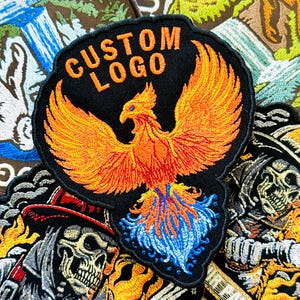May include: Embroidered patch featuring a phoenix with outstretched wings in shades of orange and yellow, with blue accents. The patch has the words "CUSTOM LOGO" in orange. Other patches with skull and floral designs are visible.