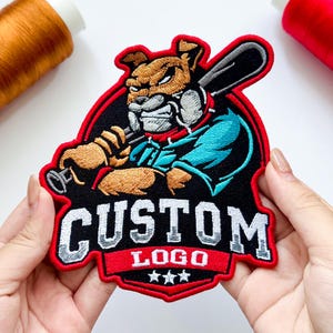 May include: An embroidered patch featuring a tan bulldog mascot holding a black baseball bat, set against a black background with a red border. The text CUSTOM LOGO is stitched in white block letters above three small white stars on a red banner.
